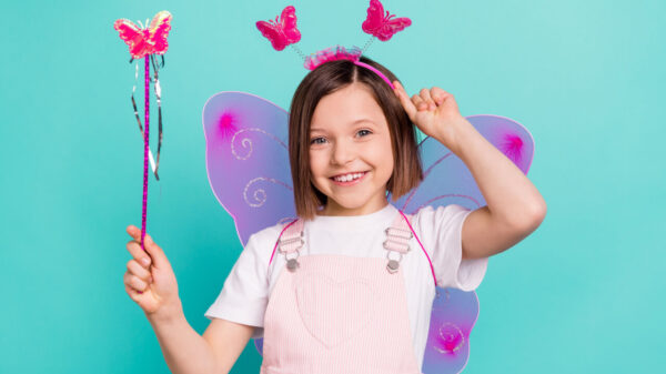 Foto de uma crianças usando asas de borboleta cor-de-rosa, sorrindo, segurando uma varinha mágica, isolada em um fundo de cor azul esverdeado.