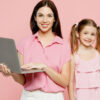 Mulher feliz, sorrindo, usando roupas casuais, ao lado de uma criança, segurando um laptop. Isoladas em um fundo rosa pastel liso.