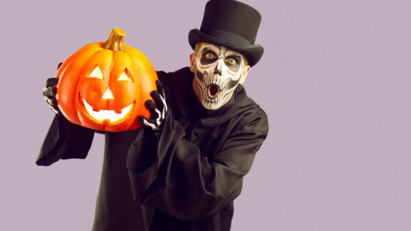 Homem usando fantasia de um esqueleto com uma capa preta e uma cartola para o Halloween. Isolado em um fundo roxo claro, segurando uma abóbora iluminada e olhando para a câmera com uma expressão surpresa no rosto.