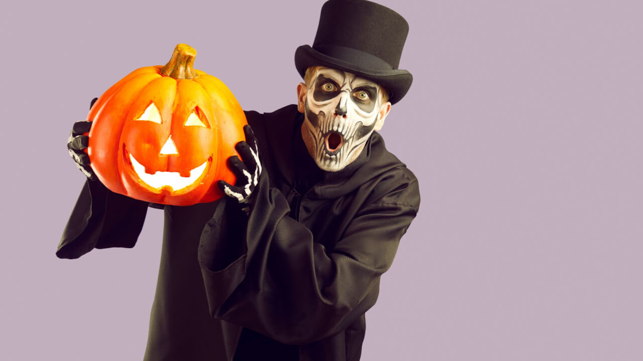 Homem usando fantasia de um esqueleto com uma capa preta e uma cartola para o Halloween. Isolado em um fundo roxo claro, segurando uma abóbora iluminada e olhando para a câmera com uma expressão surpresa no rosto.