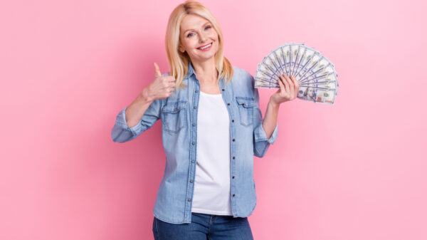 Foto de uma mulher segura notas de dinheiro, com um polegar levantado, em um fundo de cor rosa isolado.