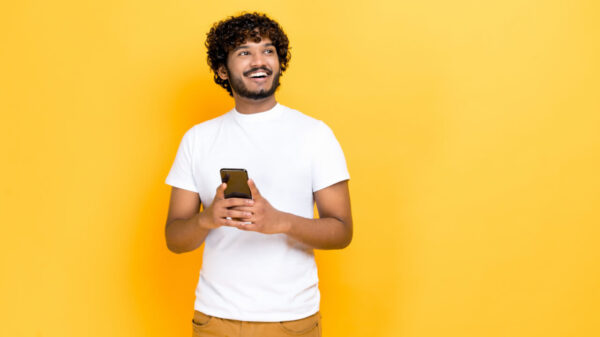Homem carismático, de camiseta branca, segurando um celular nas mãos, olhando alegremente para o lado, pensando, em pé sobre um fundo laranja isolado, sorrindo.