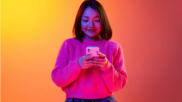 Mulher de suéter rosa usando o celular sobre um fundo laranja gradiente em luz neon. Conceito de emoções, expressão facial e inspiração.