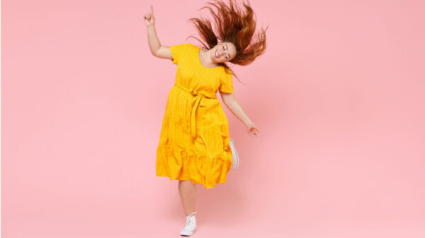 Retrato de corpo inteiro uma mulher alegre, ruiva, usando um vestido amarelo, pulando e dançando, com cabelos esvoaçantes, isolada em um estúdio de fundo de cor rosa pastel.