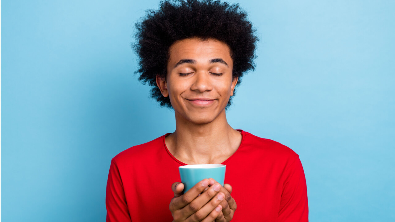 Retrato de um homem pacífico e positivo, com os olhos fechados, com as mãos segurando uma xícara de café, isolado em um fundo de cor azul.