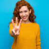 Mulher ruiva usando um suéter amarelo, feliz e fazendo o número três com os dedos.