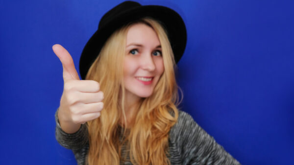 Garota sorrindo, mostrando o polegar para cima, alegre, usando um chapéu preto, sobre um fundo azul, olhando para a câmera.