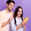 Foto de um homem e uma mulher positivos, juntos, olhando para a tela de um celular, isolados em um fundo de cor violeta.