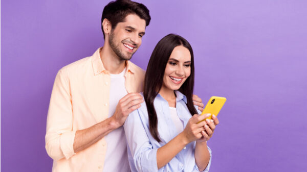 Foto de um homem e uma mulher positivos, juntos, olhando para a tela de um celular, isolados em um fundo de cor violeta.