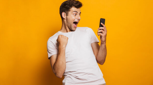Retrato de um homem animado olhando para o celular, isolado sobre um fundo amarelo, celebrando.