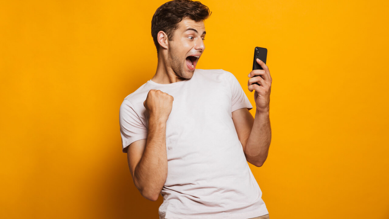 Retrato de um homem animado olhando para o celular, isolado sobre um fundo amarelo, celebrando.