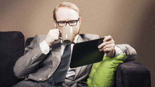 Homem sentado em um sofá, tomando um café e olhando para a tela de um tablet.