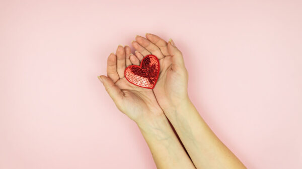 Mãos femininas segurando um coração de lantejoulas vermelhas em um fundo rosa.