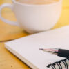 caderno, caneta e xícara branca de café em cima de uma mesa de madeira