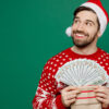 homem branco com roupas natalinas e segurando um leque de dinheiro em fundo verde