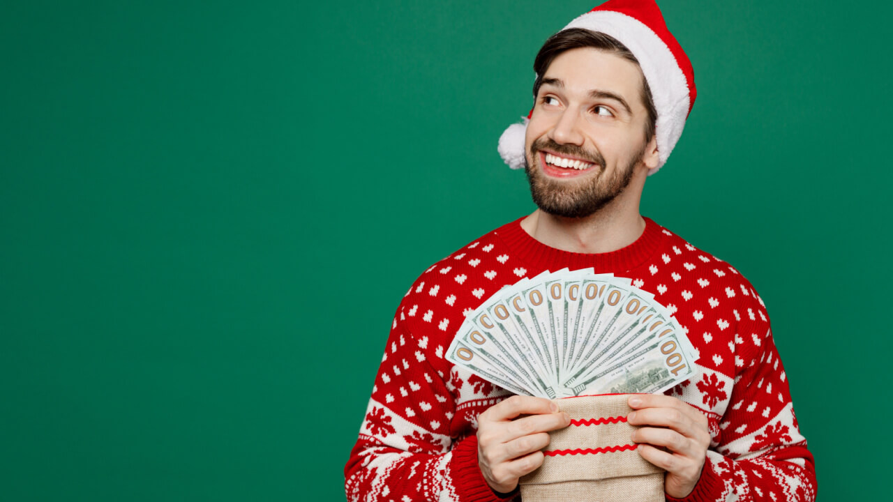 homem branco com roupas natalinas e segurando um leque de dinheiro em fundo verde