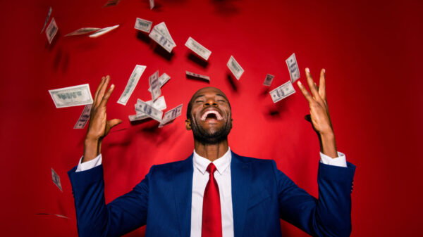 Homem usando um terno chique, elegante, moderno e azul, com os olhos fechados, sob uma chuva de dinheiro, isolado em um fundo vermelho, levantando as mãos para cima.