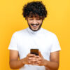 Homem de cabelos cacheados, animado e positivo, usando uma camiseta branca casual, usando celular, sorrindo, em um fundo laranja isolado.