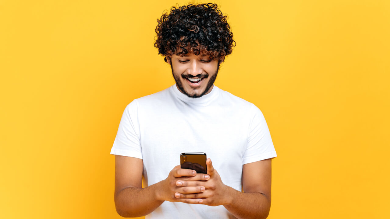 Homem de cabelos cacheados, animado e positivo, usando uma camiseta branca casual, usando celular, sorrindo, em um fundo laranja isolado.