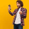 Homem, feliz, alegre, olhando para o celular em um fundo amarelo.