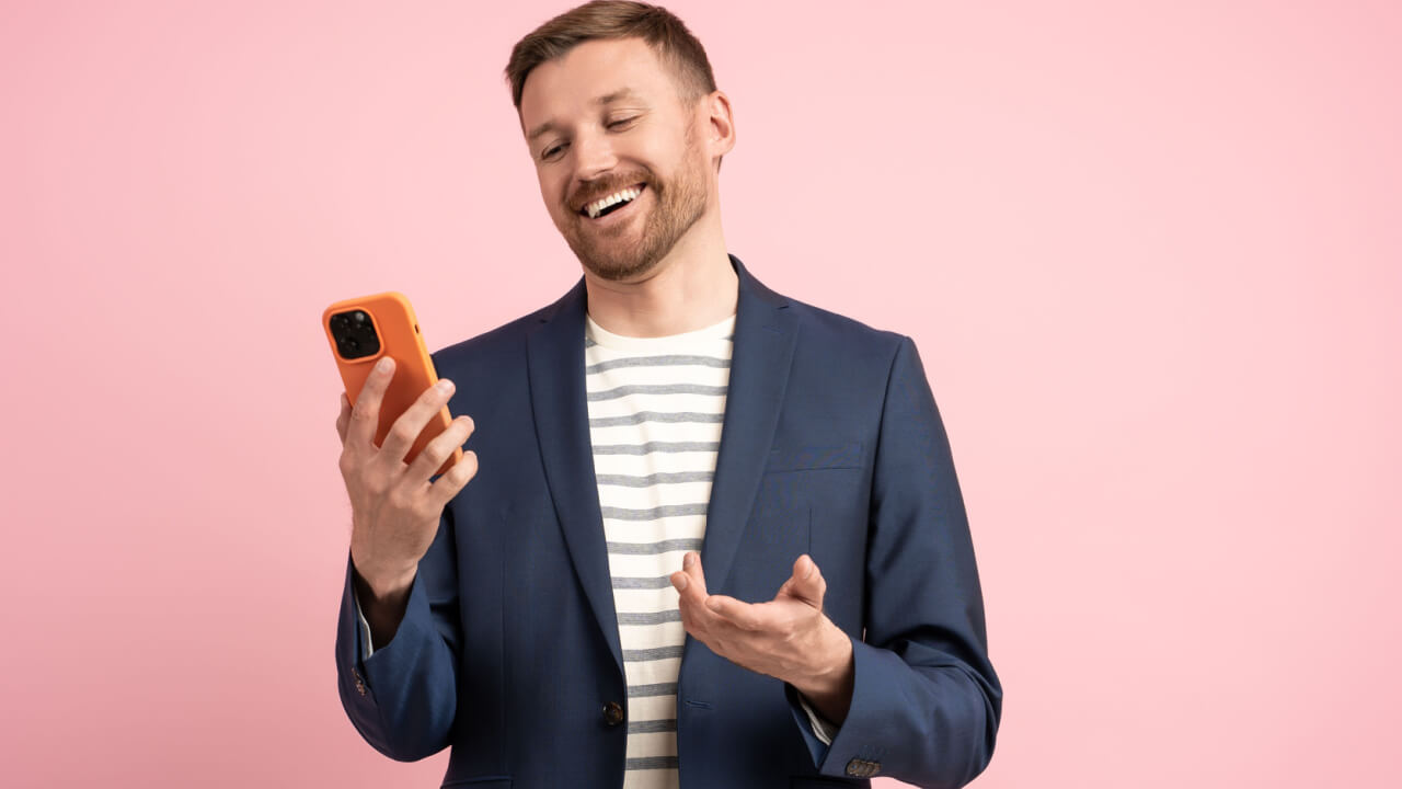 Homem sorrindo, alegre, olhando para a tela do celular, rindo, regozijando-se com algo, sobre um fundo rosa isolado.
