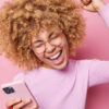 Mulher de cabelos cacheados rindo positivamente, levantando um braço, comemorando o triunfo, segurando um celular moderno, comemorando boas notícias, usando óculos transparentes, isolada sobre um fundo rosa.