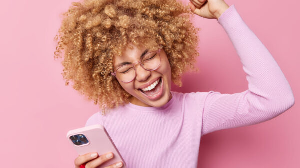 Mulher de cabelos cacheados rindo positivamente, levantando um braço, comemorando o triunfo, segurando um celular moderno, comemorando boas notícias, usando óculos transparentes, isolada sobre um fundo rosa.