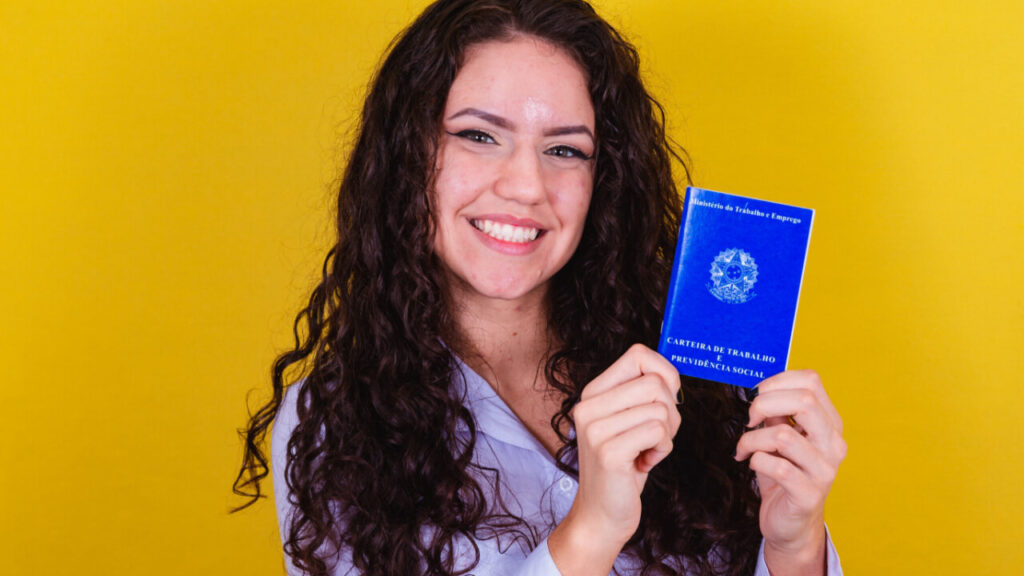 mulher sorrindo segurando uma carteira de trabalho em fundo amarelo