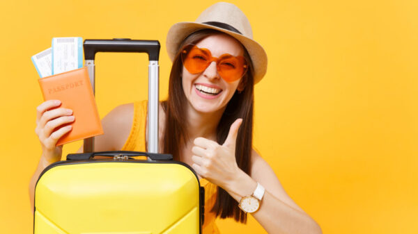 Mulher turista usando roupas casuais de verão, com um chapéu e uma mala, mostrando polegar para cima, isolada em um fundo amarelo. Conceito de viagem de voo aéreo.