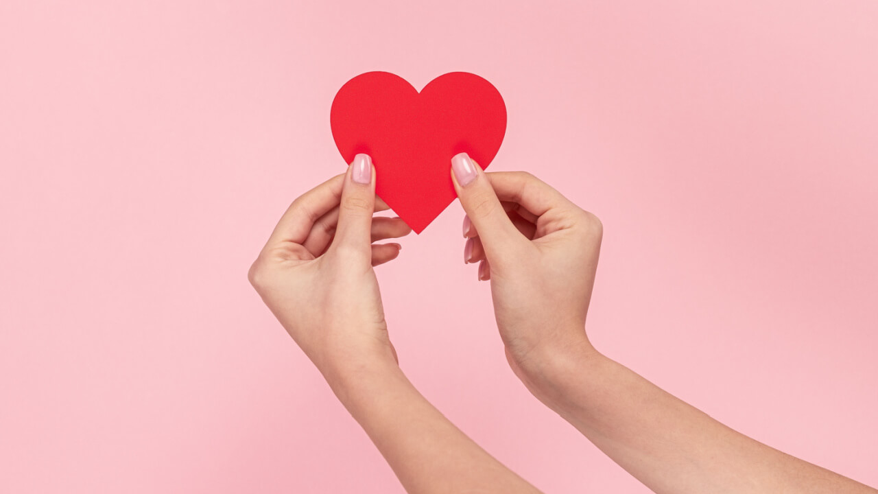 Mãos femininas segurando delicadamente um coração vermelho de papel contra um fundo rosa.