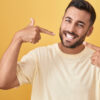 homem branco sorrindo com os dedos indicadores apontando o sorriso em fundo amarelo