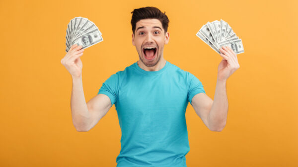 homem branco vestindo camiseta azul e segurando dois leques de dinheiro, um em cada mão, em fundo laranja