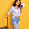 Retrato de corpo inteiro de uma mulher feliz, segurando passagens, um passaporte e uma mala, posando em um fundo amarelo, indo fazer um passeio.