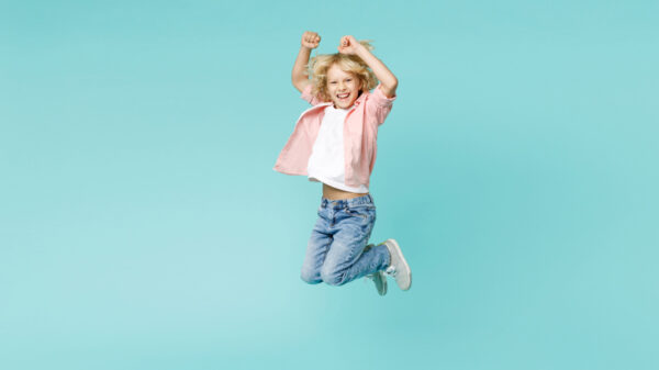 Foto de corpo todo de um menino feliz, pulando, fazendo gesto de vencedor, isolado em um fundo de cor azul turquesa em um estúdio.
