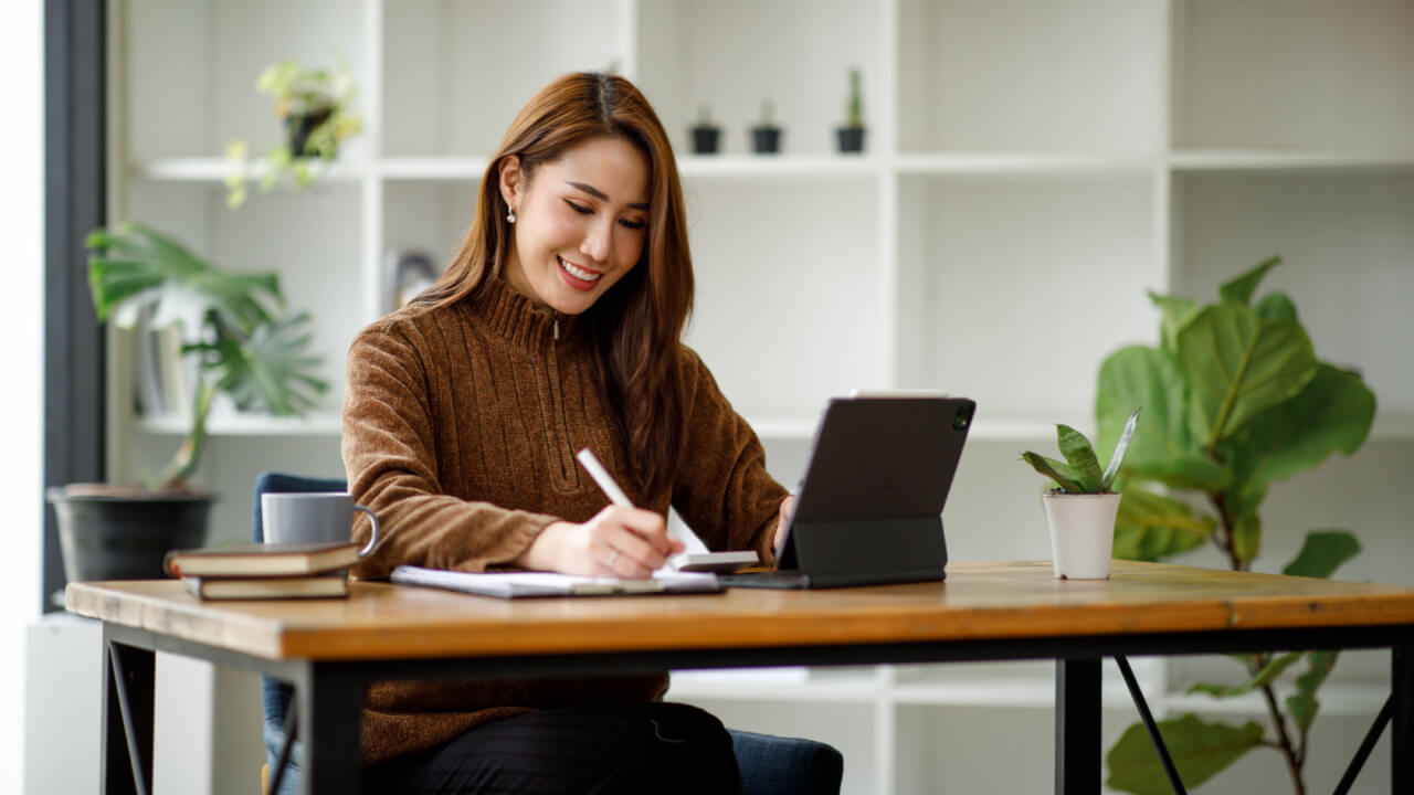 Mulher com traços asiáticos trabalhando com caderno e tablet em uma escritório de estantes brancas