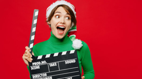 Mulher surpresa, usando uma blusa verde de gola alta e um gorro de Papai Noel, posando, segurando uma claquete, olhando de lado, isolada em um fundo vermelho liso. Conceito natal.