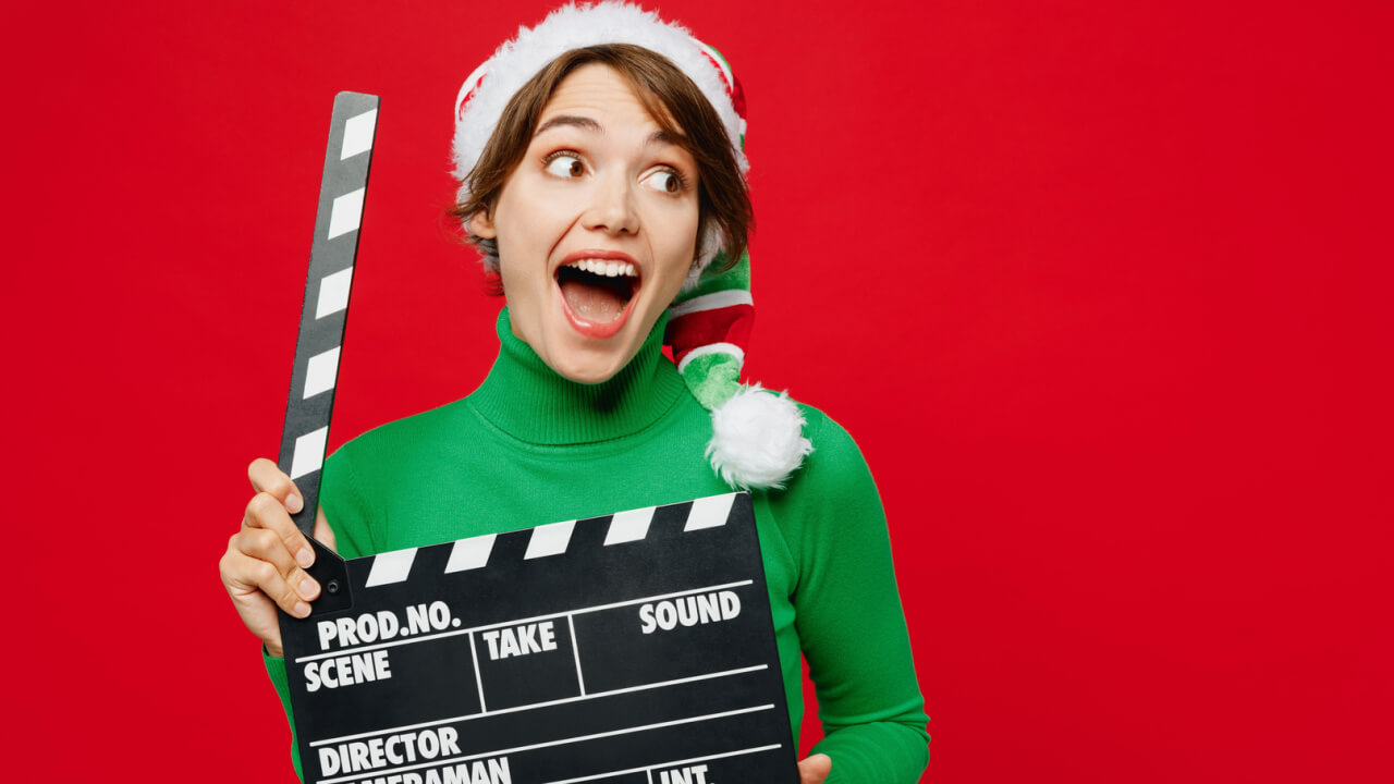 Mulher surpresa, usando uma blusa verde de gola alta e um gorro de Papai Noel, posando, segurando uma claquete, olhando de lado, isolada em um fundo vermelho liso. Conceito natal.