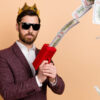 Foto de um homem rico, com uma coroa na cabeça e com dinheiro, isolado em um fundo de cor bege.