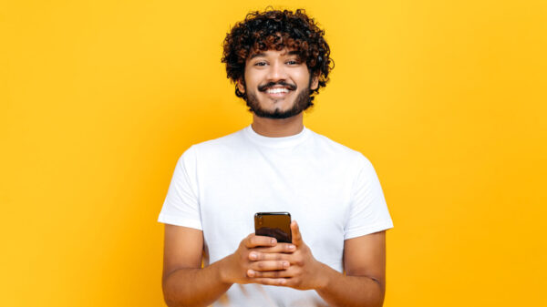 Homem positivo, usando um celular, navegando na Internet, parado em um fundo amarelo isolado, olhando para a câmera, sorrindo.