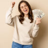 Mulher entusiasmada, segurando notas de dinheiro, celebrando e gritando de alegria, contra um fundo bege.