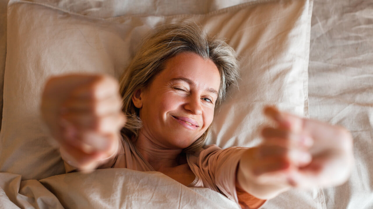 mulher loira deitada em um travesseiro de lençol marrom claro com os braços esticados para cima assim ficando desfocados na foto