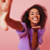 mulher negra brincando com a camera vestindo roxo e em fundo rosa