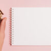 à direta há um caderno em folha em branco e à esquerda uma casinha de madeira segurada em uma mão em fundo rosa