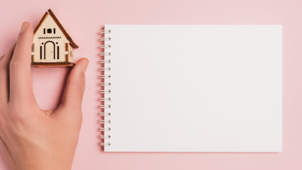 à direta há um caderno em folha em branco e à esquerda uma casinha de madeira segurada em uma mão em fundo rosa