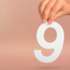 mão segurando um número 9 branco em fundo rosa