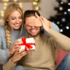 mulher abraçando um homem e colocando a mão direita em seu olho para entregar um presente em fundo de decoração natalina desfocada