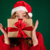 Mulher com gorro de Papai Noel, cobrindo o rosto com uma caixa de presente, isolada em um fundo verde, usando um suéter.