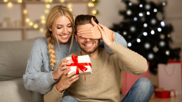 mulher abraçando um homem e colocando a mão direita em seu olho para entregar um presente em fundo de decoração natalina desfocada