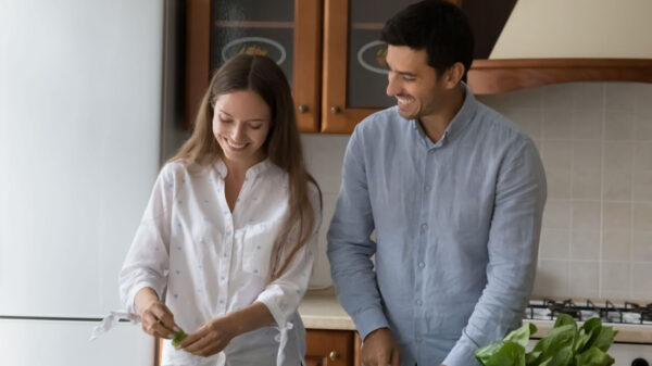 casal feliz temperando salada na cozinha de casa com armários de madeira