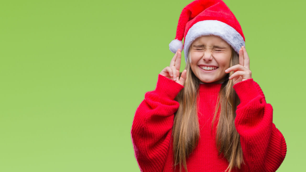 no canto direito da imagem tem uma menina loira de cabelos lisos usando roupa vermelha e chapéu de natal e cruzando os dedos em fundo verde